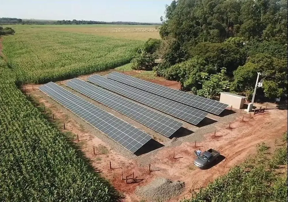 Vista aérea de uma instalação de painéis solares em uma área rural, cercada por plantações e vegetação, com uma caminhonete estacionada próxima