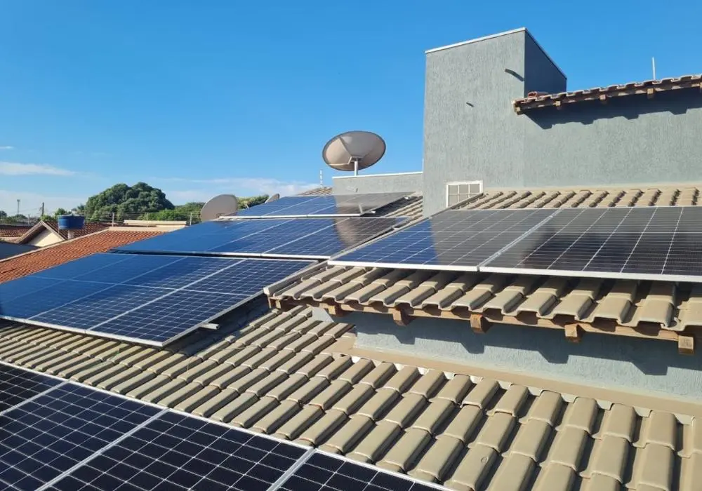 Painéis solares instalados em um telhado de telhas em uma área residencial, com antenas parabólicas visíveis e céu azul ao fundo