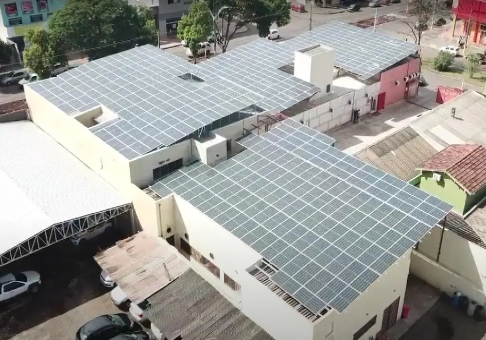 Vista aérea de uma instalação de painéis solares cobrindo o telhado de um complexo de edifícios comerciais, com árvores e carros visíveis ao redor