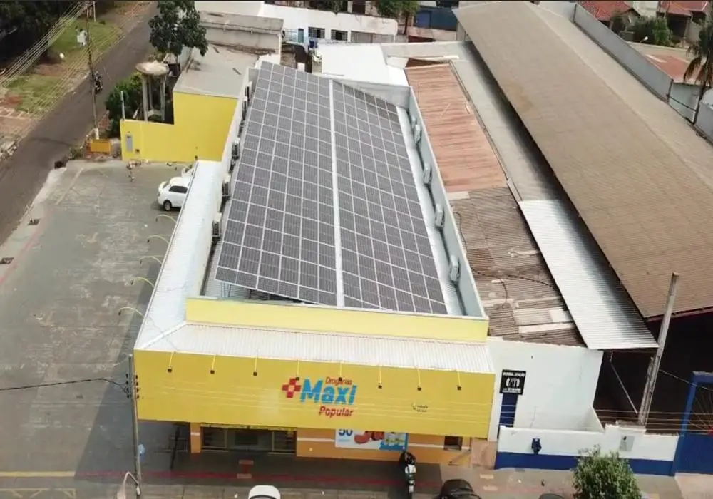 Vista aérea de uma instalação de painéis solares no telhado de um edifício comercial amarelo com o nome 'Maxi Popular', localizado em uma área urbana